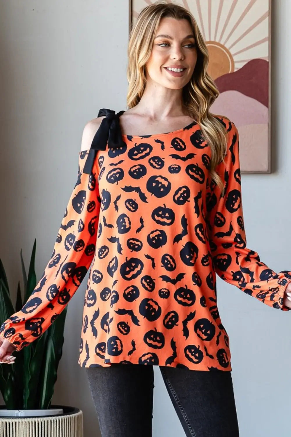 Woman wearing Heimish pumpkin print one-shoulder t-shirt with black bow detail on shoulder, styled for fall Halloween season.