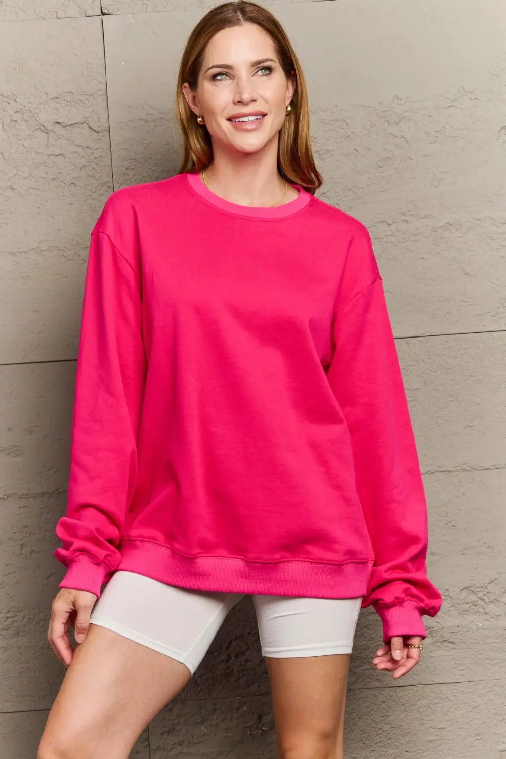 Woman wearing bright pink full size round neck long sleeve sweatshirt standing against textured wall