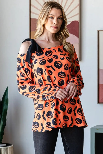 Woman wearing an orange Heimish pumpkin print one-shoulder t-shirt with black bow detail, paired with dark jeans indoors.