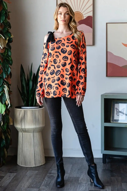 Woman wearing Heimish orange pumpkin print one-shoulder t-shirt paired with black jeans and ankle boots indoors