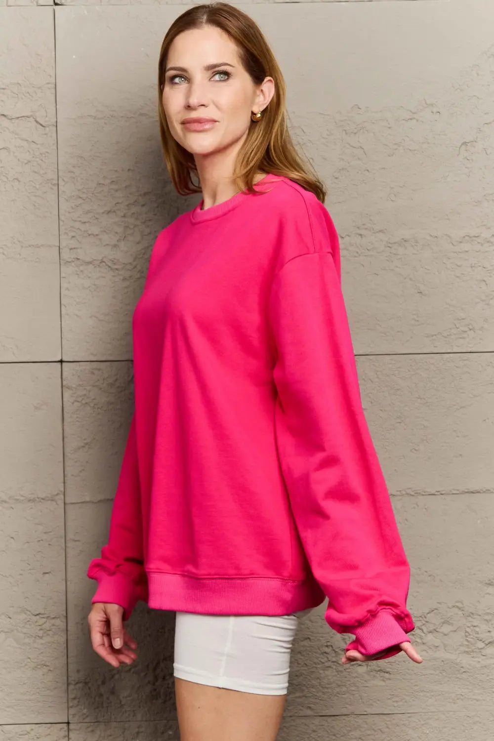 Woman wearing a bright pink full size round neck long sleeve sweatshirt standing against a textured wall