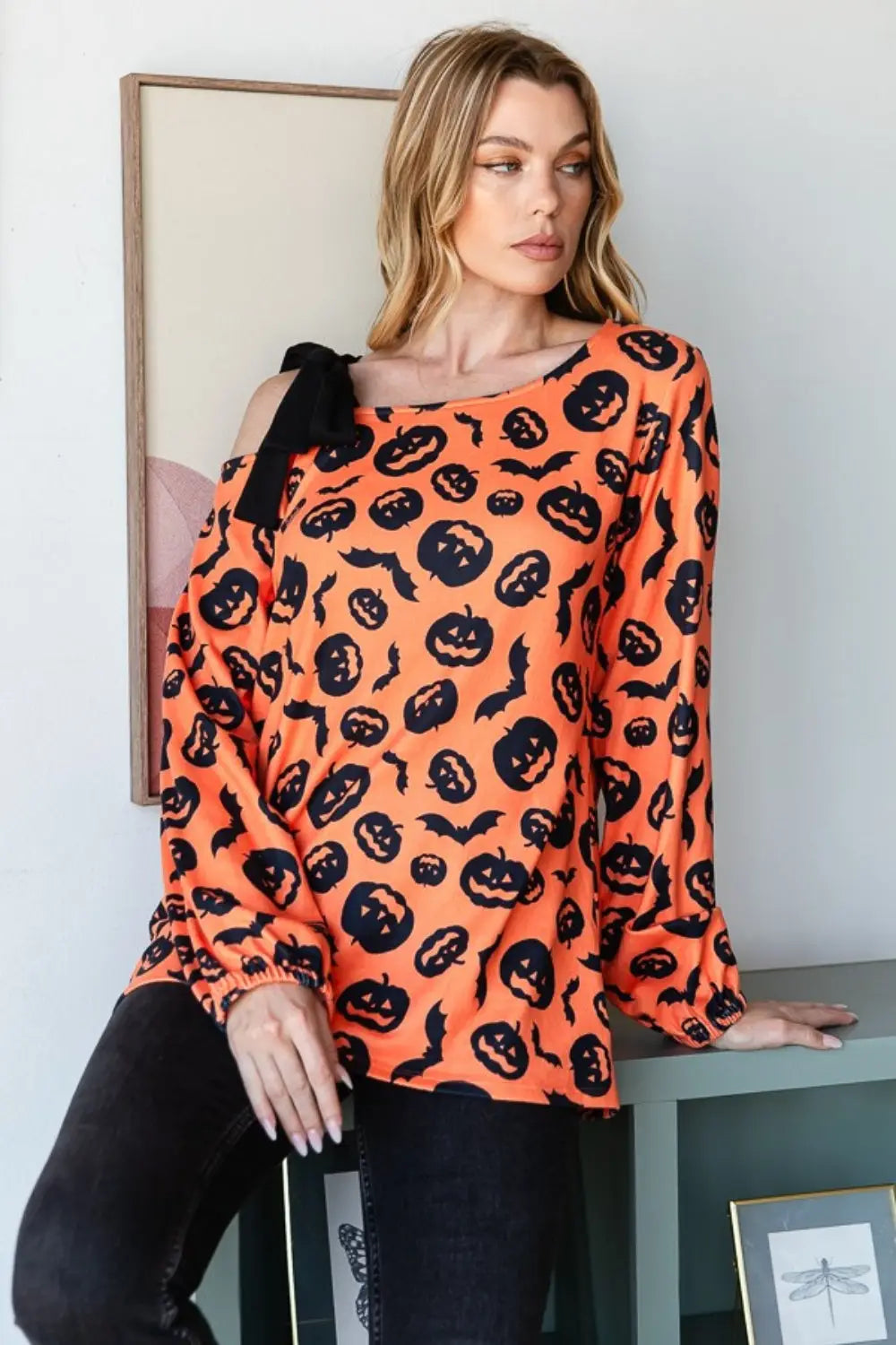 Woman wearing Heimish Halloween pumpkin print one-shoulder orange t-shirt with black bow detail sitting by green table