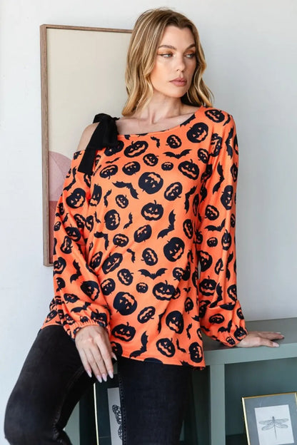 Woman wearing Heimish Halloween pumpkin print one-shoulder orange t-shirt with black bow detail sitting by green table