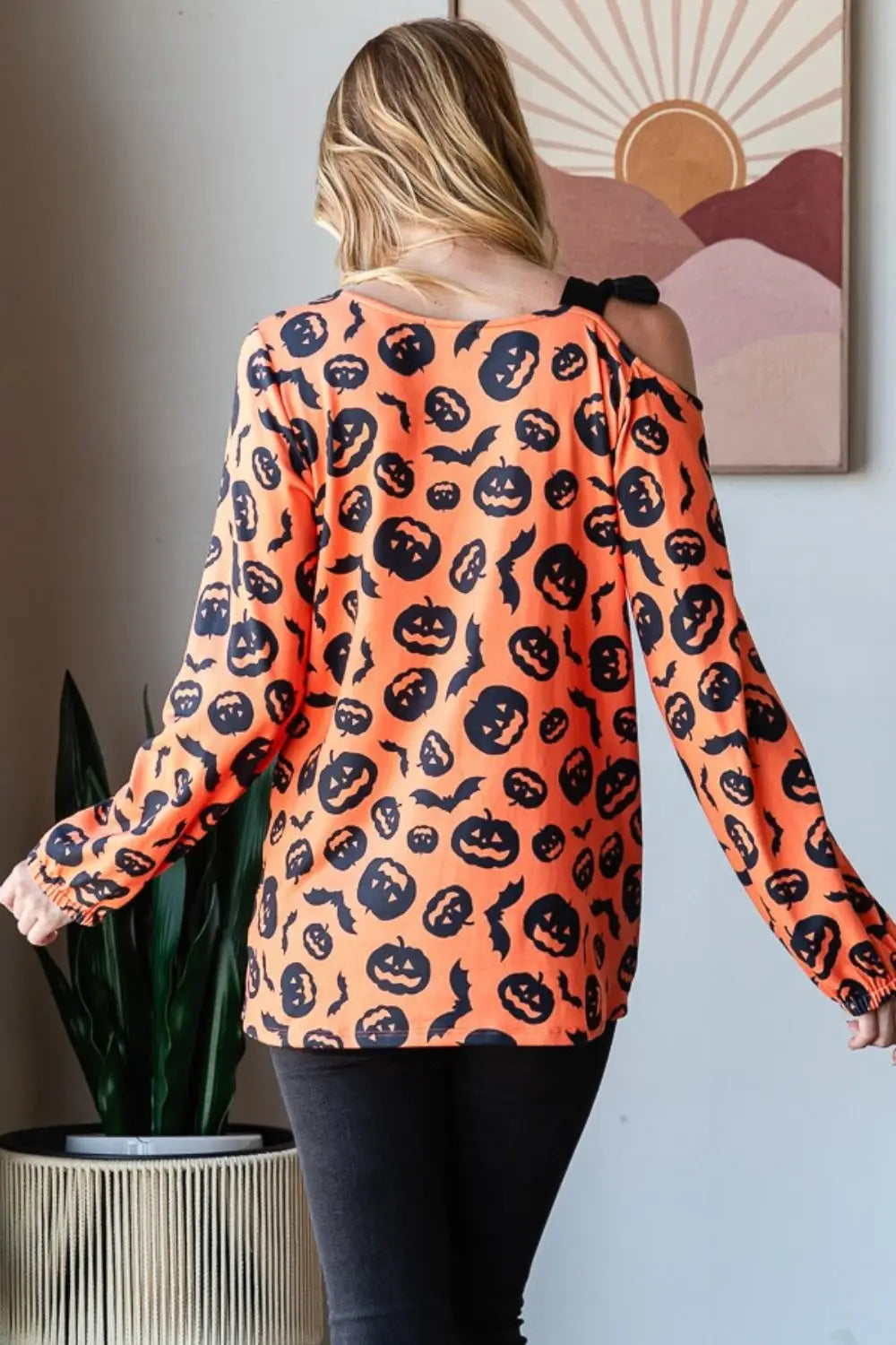 Woman wearing Heimish orange one-shoulder t-shirt with black pumpkin and bat prints standing indoors